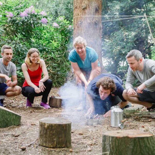 A group surrounding a man in the woods watching him blowing a Bushcraft fire