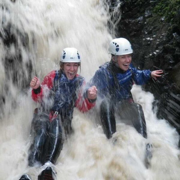 Two ladies wearing wetsuits and helmets Canyoning under a waterfall