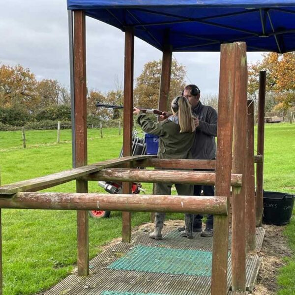 A girl dressed in a green boiler suit, being assisted by an instructor on Clay Pigeon Shooting
