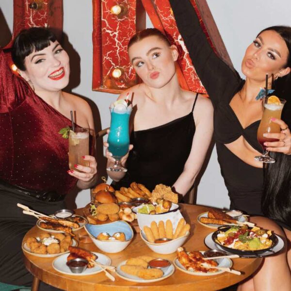 Three women sat at a table filled with Grazer dishes in Revolution holding their cocktails in the air