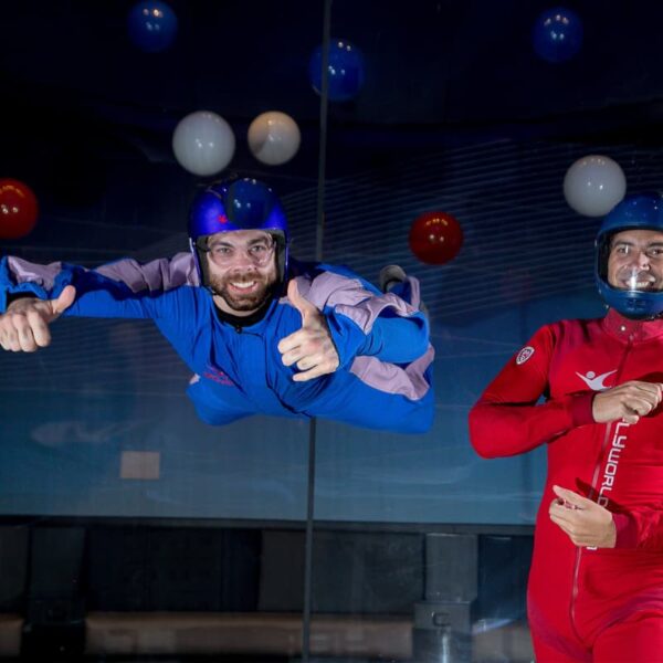 Man with a beard smiling, putting a thumbs up whilst indoor skydiving next to an instructor in a red boiler suit