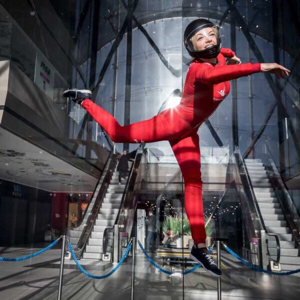 A woman dressed in a jumpsuit and helmet mid air doing Indoor Skydiving