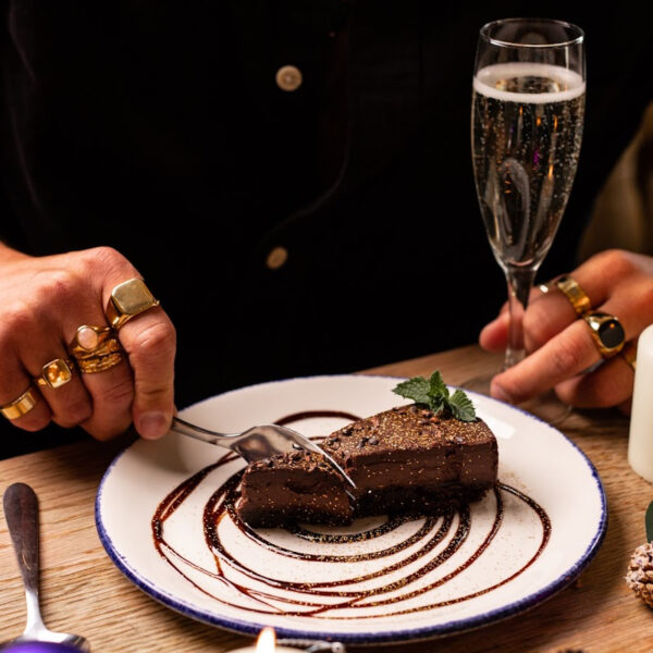 Person cutting into a slice of chocolate brownie drizzled with chocolate sauce and holding a glass of prosecco.