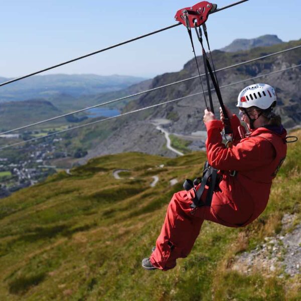 A lady wearing a red jumpsuit and helmet, flying through the sky on the Zip Wire Titan